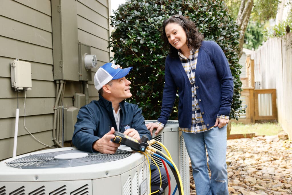 An HVAC technician testing an outdoor AC unit next to a gray-green home while speaking with the homeowner. Tall bush behind them.