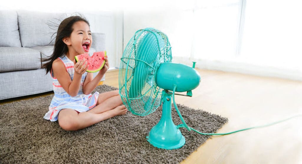 Young girl holding a slice of watermelon sitting in front of a blue fan on the floor in a living room.