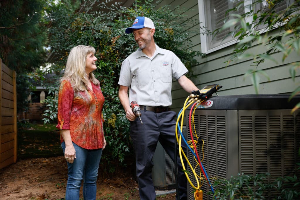 HVAC technician testing an outdoor AC unit next to a green home while speaking with the homeowner. Tall bush behind them.