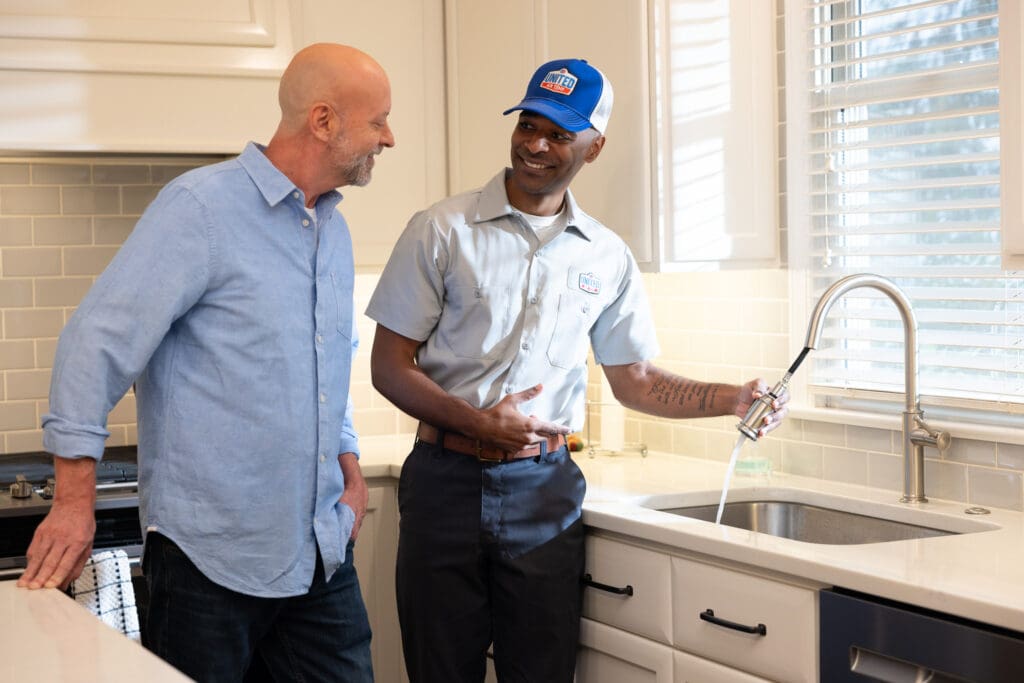 Technician explaining plumbing service to a homeowner while standing in the update white kitchen, holding the sprayer faucet of the kitchen sink.