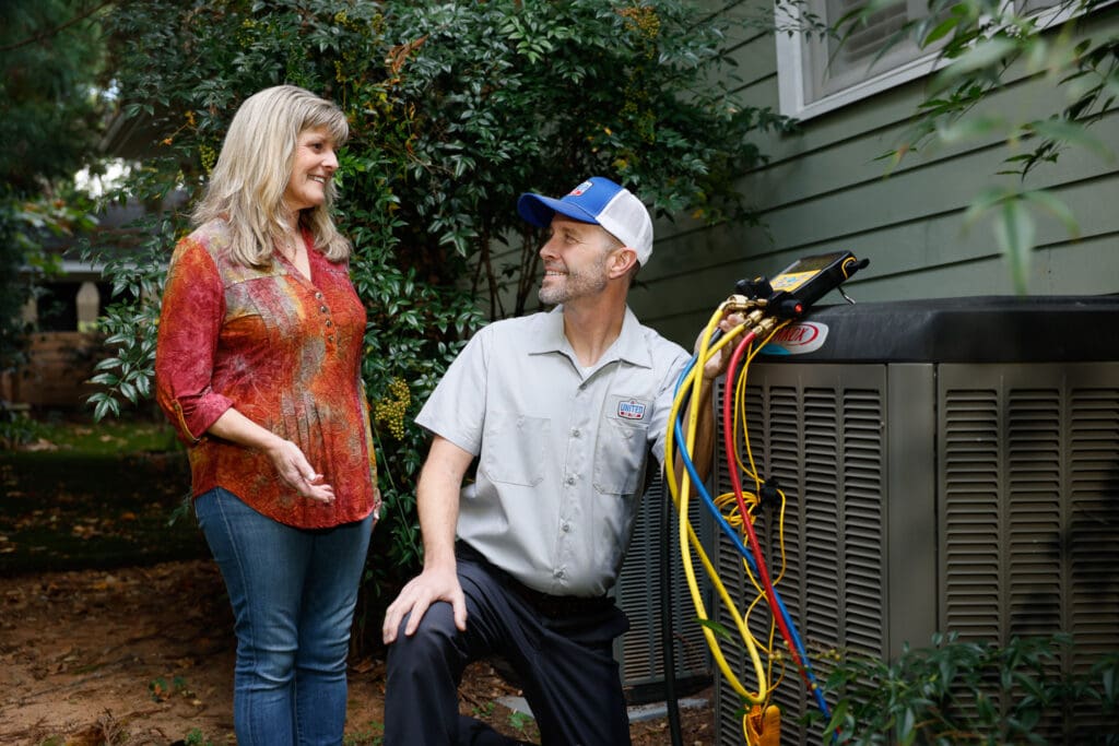 An HVAC technician testing and outdoor AC unit next to a green home while talking with homeowner. Tall bush behind them.