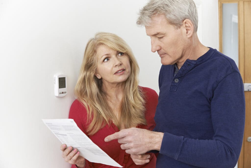 Concerned couple with utility bill in front of a thermostat on a white wall.